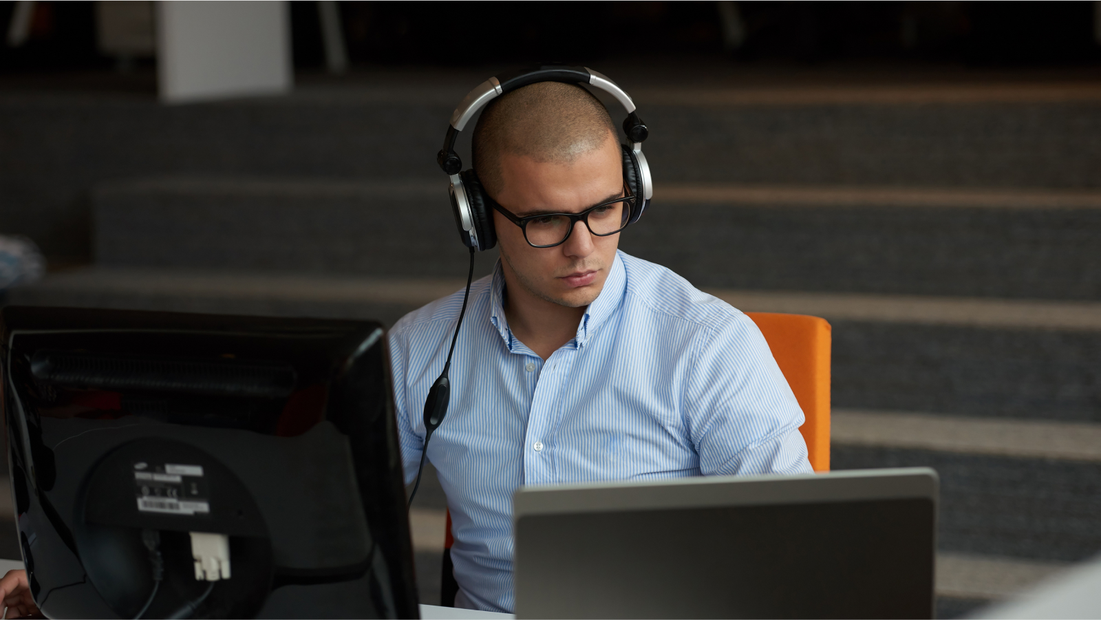 The image shows a man sitting in front of a computer. He is wearing a blue, striped long-sleeve shirt. The man has large, black glasses and is wearing headphones. 