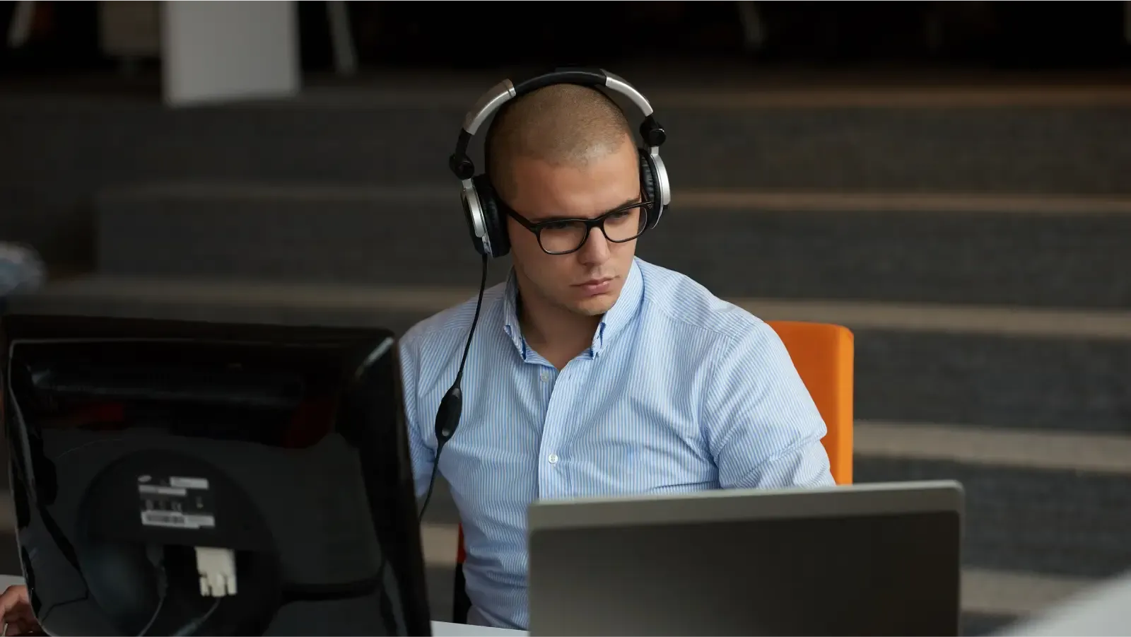 The image shows a man sitting in front of a computer. He is wearing a blue, striped long-sleeve shirt. The man has large, black glasses and is wearing headphones. 