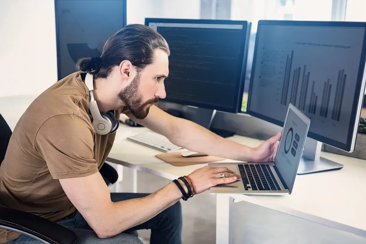 The picture presents a man sitting at the desk and staring at the laptop screen. Charts and a code editor are displayed on the laptop screen and on the other two monitors standing on the desk