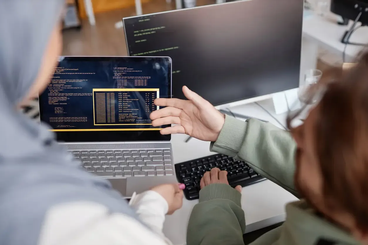 The picture shows two people sitting at the computers. One person explains something to the other and pointing on her monitor with his hand