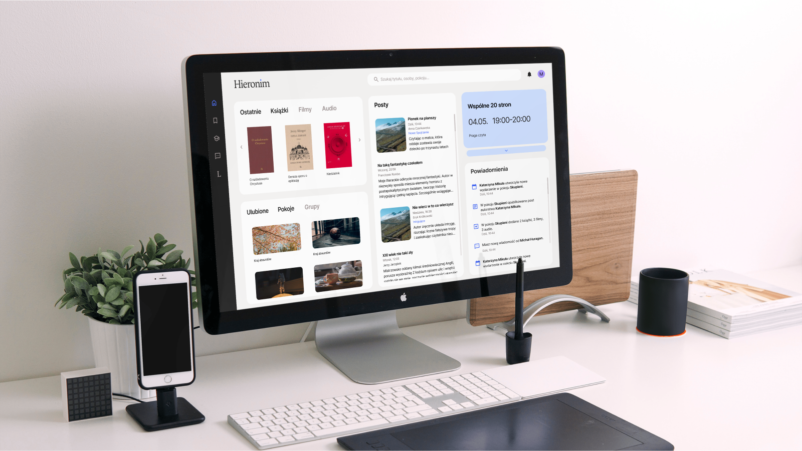 Ekran komputera na biurku z wyświetlonym projektem strony głównej użytkownika biblioteki multimedialnej Hieronim. Pokazane są ostatnie książki, filmy, audiobooki, ulubione grupy, feed z postam z grup, kalendarz wydarzeń grup, oraz powiadomienia z ostatnimi wydarzeniami w serwisie. Wygląd jest jasny, schludny, zaokrąglone panele mają zakładki . Delikatnie wyróżnią się z tła, ułatwiając skanowanie strony.