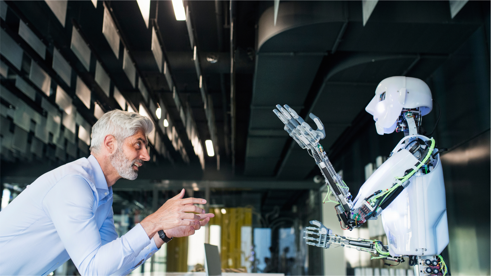An elderly man with gray hair and a beard, wearing a light blue shirt, is interacting with a humanoid robot. The robot, which has a white, streamlined body with visible mechanical components and wiring, raises its hands as if mirroring the man's gestures.