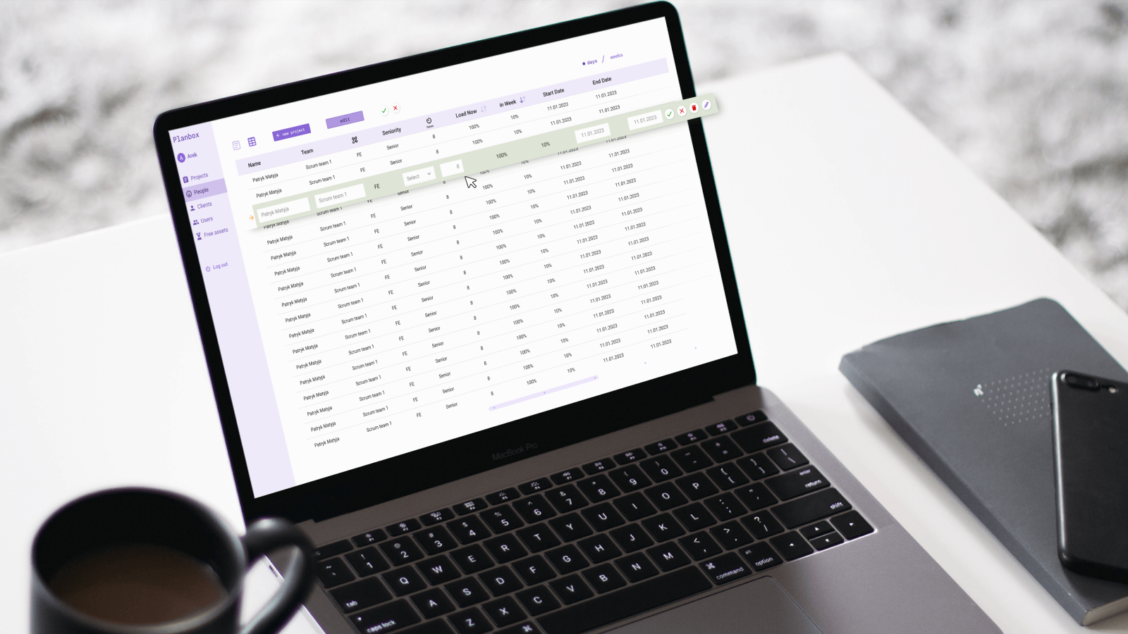 Laptop on a desk with the Planbox application open, displaying a list of tasks and people assigned to various projects. In the foreground, there is a cup of coffee, and next to it a notebook and a phone.