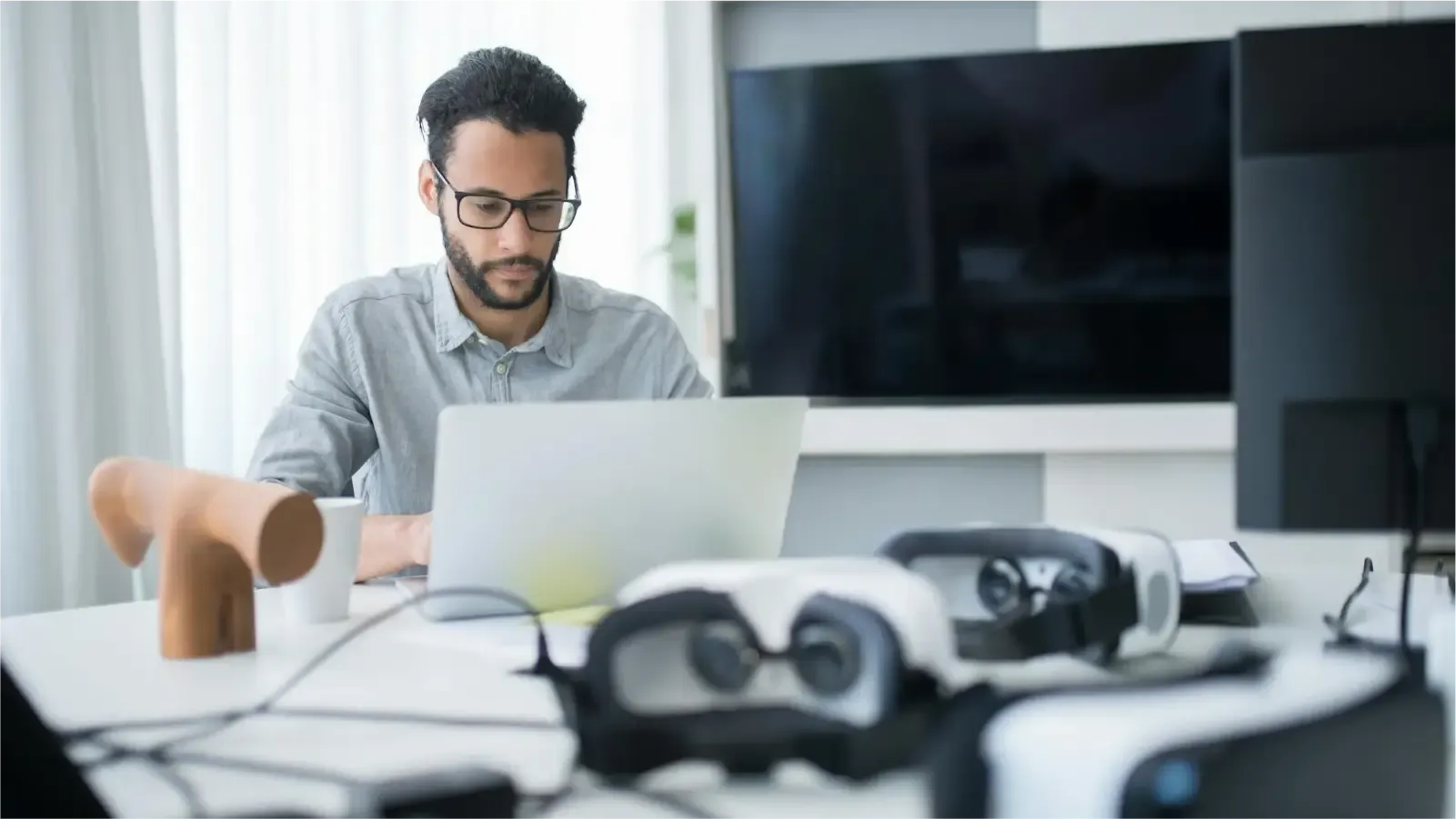 A man with short, dark curly hair and a beard works on a laptop in a modern office. He wears glasses and a light gray shirt. On the table are two VR headsets, a white coffee cup, and a wooden sculpture. A large flat-screen TV and part of a computer monitor are visible in the background. Natural light comes through sheer white curtains.